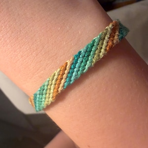 May include: A woven friendship bracelet in shades of teal, yellow, and orange. The bracelet is made of braided threads and is worn on a wrist. The colors are arranged in diagonal stripes.