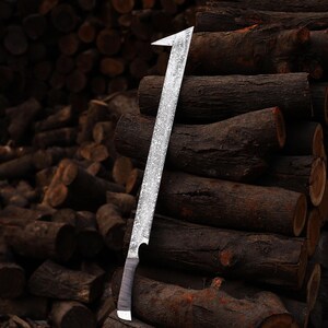 May include: A large, silver machete with a textured blade and a dark handle. The machete is leaning against a stack of brown logs, with more logs in the background. The blade has a sharp, angled tip.