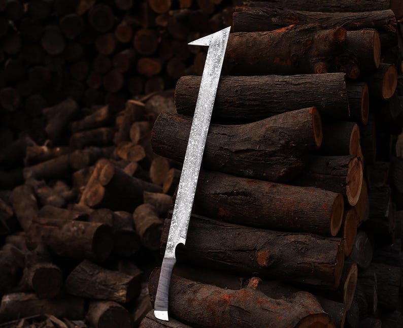 May include: A large, silver machete with a textured blade and a dark handle. The machete is leaning against a stack of brown logs, with more logs in the background. The blade has a sharp, angled tip.