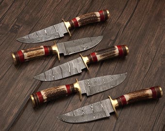 May include: Five hunting knives with patterned blades and antler handles. The handles have red and gold accents. The knives are arranged on a dark wood surface, showcasing their craftsmanship and design.