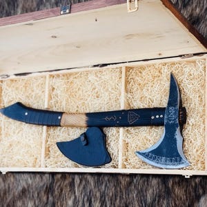 Può includere: Un'ascia con manico in legno naturale e nero e una lama curva con simboli runici incisi e un emblema Valknut. L'ascia include un fodero in pelle nera ed è presentata all'interno di una scatola di legno rettangolare riempita di trucioli.