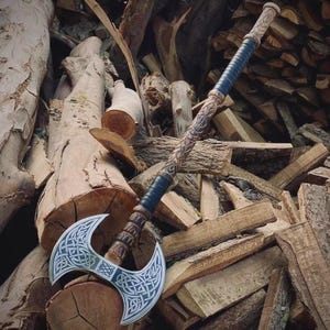 May include: A double-bitted battle axe featuring a silver-colored metal head with intricate Celtic knotwork engravings. The long wooden handle is carved with decorative patterns and includes two sections of dark blue leather wrapping.