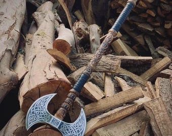 May include: A double-bitted battle axe featuring a silver-colored metal head with intricate Celtic knotwork engravings. The long wooden handle is carved with decorative patterns and includes two sections of dark blue leather wrapping.
