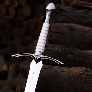 Peut inclure: Gros plan d'une épée argentée et blanche avec une lame polie. Le manche est blanc avec un motif nervuré. La garde transversale a un motif décoratif avec une gemme bleue. L'épée repose sur une pile de bûches brun foncé.