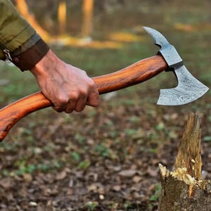 May include: A handcrafted axe with a wooden handle and a patterned steel blade. The axe is held in a hand, ready for use. The handle is a warm brown colour, and the blade has a unique, textured design. The axe is designed for chopping wood.