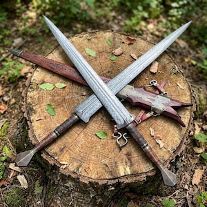 May include: Two double-edged swords with intricate etched patterns on the steel blades, dark brown leather-wrapped grips, and diamond-shaped pommels. A matching dark wood scabbard with metallic accents and leather straps lies beneath the crossed blades.