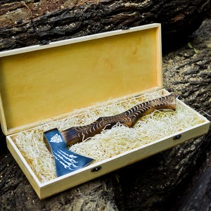 May include: A wooden box with a light interior contains a handcrafted axe. The axe has a dark wooden handle with detailed carvings and a blue-grey blade with a white design. The box is lined with straw.