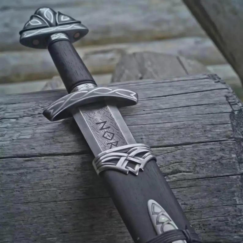 May include: A Viking sword with a dark wooden handle and silver-toned fittings. The blade features an etched design with the word "NOR". The sword is sheathed in a dark scabbard, resting on a wooden surface. The design suggests a historical or fantasy theme.