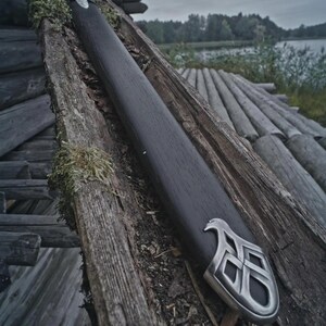 May include: A black sword in a wooden scabbard with a silver decorative end. The sword rests on weathered wooden planks with moss. The scene is set outdoors with a lake in the background.