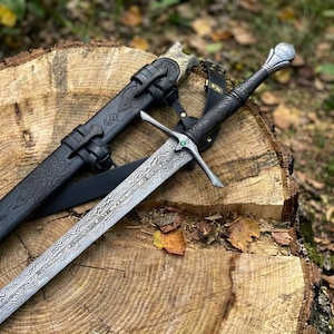 Peut inclure: Une épée en acier avec une lame à motifs et une poignée noire ornée d'une petite pierre verte sur la garde. L'épée est accompagnée d'un fourreau en cuir noir avec des motifs gaufrés complexes et des lanières en cuir noir pour la fixation.