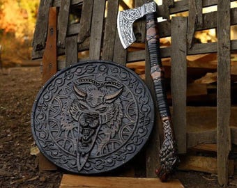 May include: A Viking-style axe and shield set. The axe features a silver blade with intricate designs and a wrapped handle. The shield is round, dark-colored, and carved with a wolf's head.