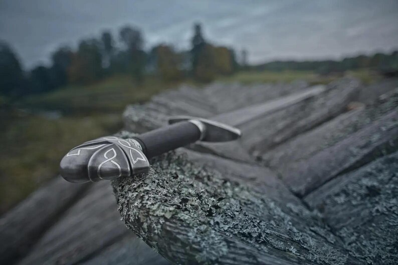 May include: Close-up of a sword with a silver hilt and a dark handle, lying on a weathered, grey wooden surface. The background is blurred, showing a natural outdoor setting with trees and a cloudy sky.