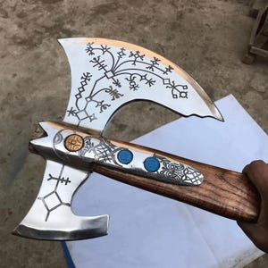 May include: A double-bitted axe with a polished silver metal head featuring intricate black etched runic patterns. The handle is made of brown wood, and the metal head includes a circular gold-toned accent and two small blue circular inlays.