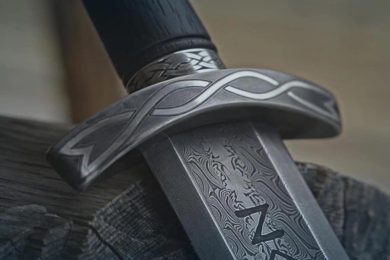 May include: Close-up of a detailed sword with a patterned blade and handle. The metalwork features intricate designs and a dark, textured handle. The sword's guard has a decorative, curved shape.