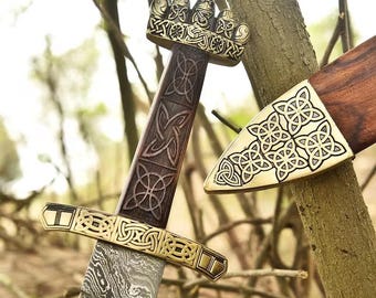 May include: A detailed image of a sword with a patterned blade and intricate gold-colored metalwork on the hilt and guard. The wooden handle features carved designs. The sword is partially sheathed in a wooden scabbard with matching metal accents.