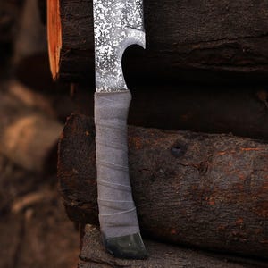May include: A close-up shot of a handmade knife featuring a textured blade with a speckled pattern and a grey, wrapped handle. The blade has a curved design. The knife is set against a backdrop of stacked, weathered wooden logs, creating a rustic aesthetic.