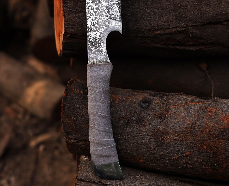 May include: A close-up shot of a handmade knife featuring a textured blade with a speckled pattern and a grey, wrapped handle. The blade has a curved design. The knife is set against a backdrop of stacked, weathered wooden logs, creating a rustic aesthetic.
