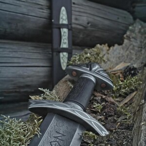 May include: A close-up of a sword with an intricate blade design and a decorative hilt, set against a backdrop of moss and wood. The sword features a dark handle and a detailed guard. A matching sheath is also visible.