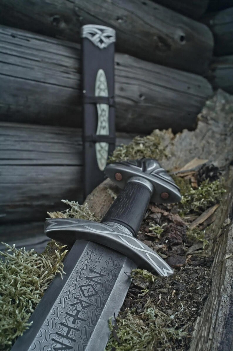 May include: A close-up of a sword with an intricate blade design and a decorative hilt, set against a backdrop of moss and wood. The sword features a dark handle and a detailed guard. A matching sheath is also visible.
