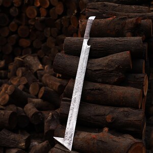 May include: A silver sword with an unusual angular blade, set against a background of stacked logs. The blade has a textured finish, and the handle is wrapped in a light-coloured material. The sword's distinctive shape and the rustic setting create a striking visual.