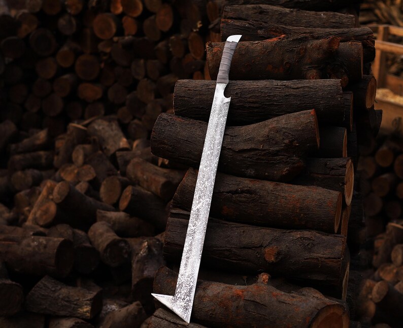 May include: A silver sword with an unusual angular blade, set against a background of stacked logs. The blade has a textured finish, and the handle is wrapped in a light-coloured material. The sword's distinctive shape and the rustic setting create a striking visual.