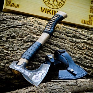 May include: A Viking axe with a wooden handle bound in black leather. The silver axe head features etched designs. A black leather sheath is included. The axe is displayed on a textured wooden surface.