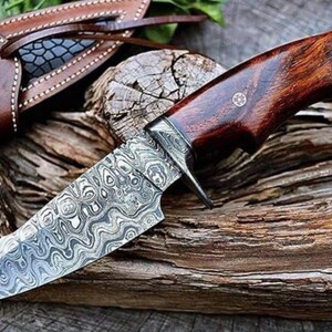 May include: A hunting knife with a patterned steel blade and a dark wood handle. The knife has a full tang construction and comes with a brown leather sheath. The blade features a distinctive, swirling pattern.