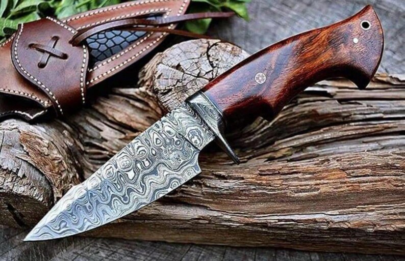 May include: A hunting knife with a patterned steel blade and a dark wood handle. The knife has a full tang construction and comes with a brown leather sheath. The blade features a distinctive, swirling pattern.