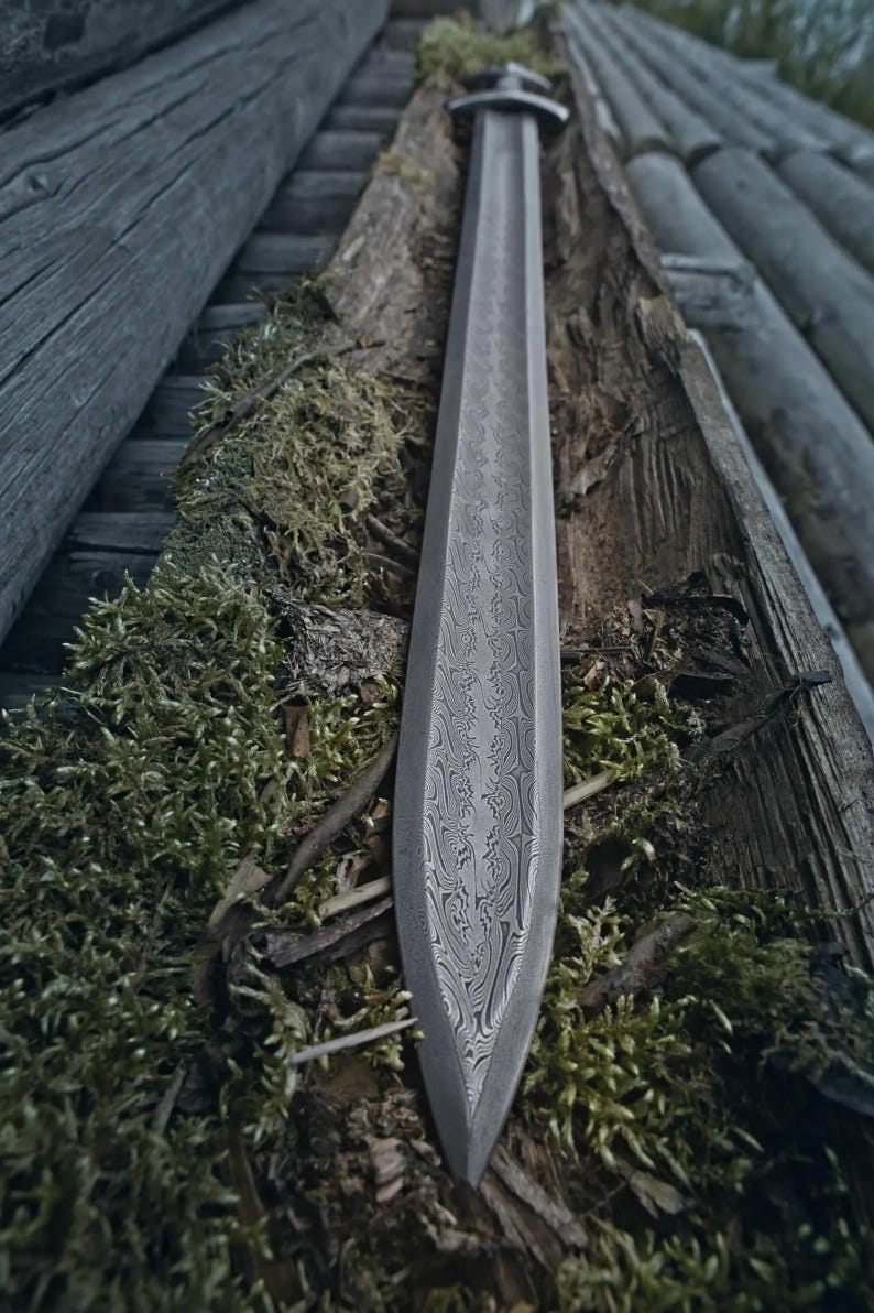 May include: A long, grey sword with a patterned blade lies on moss and wood. The blade has a detailed, swirling design. The handle is visible at the top, and the tip of the blade points downwards.