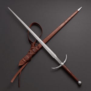 May include: A medieval-style sword with a long, silver blade and a brown leather-wrapped handle. The sword is paired with a brown leather sheath and a leather strap. The design features a crossguard and a pommel.