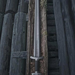 May include: A silver sword with the word "NORTHMEN" etched on the blade, set against a backdrop of weathered wooden planks. The sword features a detailed hilt and a dark handle, evoking a sense of history and craftsmanship.
