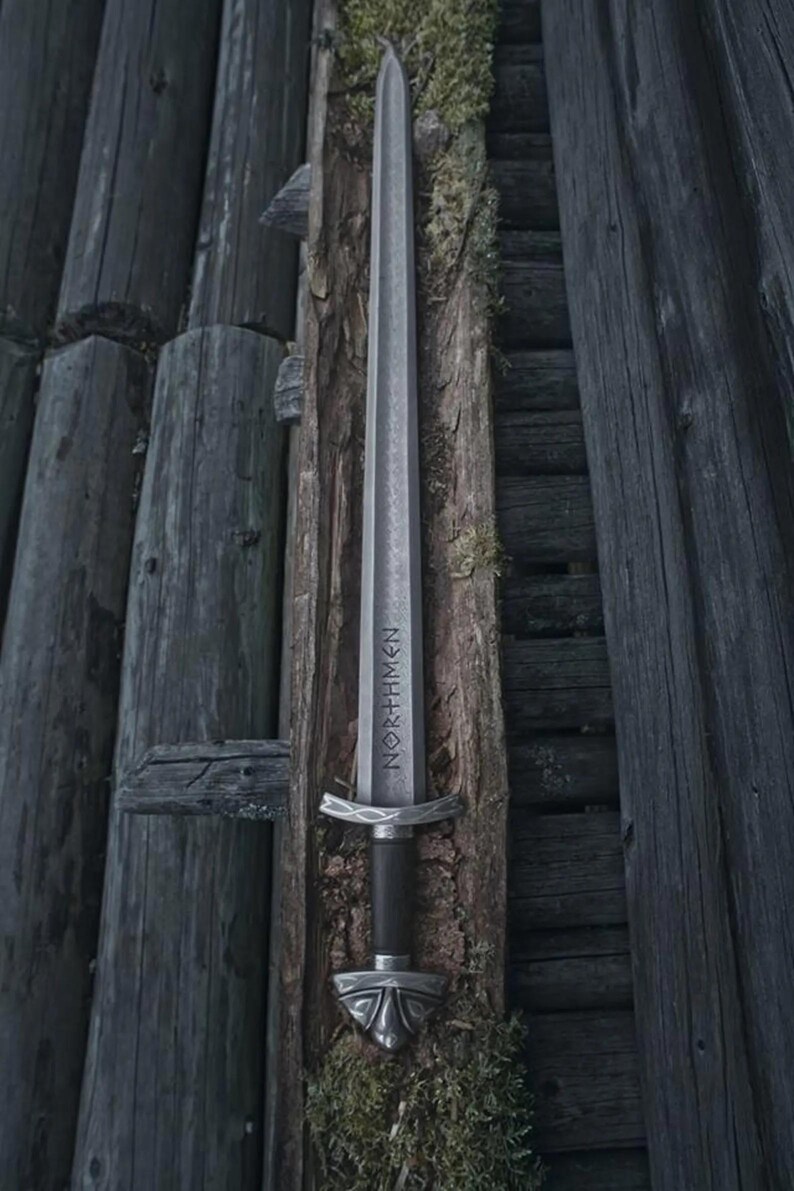 May include: A silver sword with the word "NORTHMEN" etched on the blade, set against a backdrop of weathered wooden planks. The sword features a detailed hilt and a dark handle, evoking a sense of history and craftsmanship.