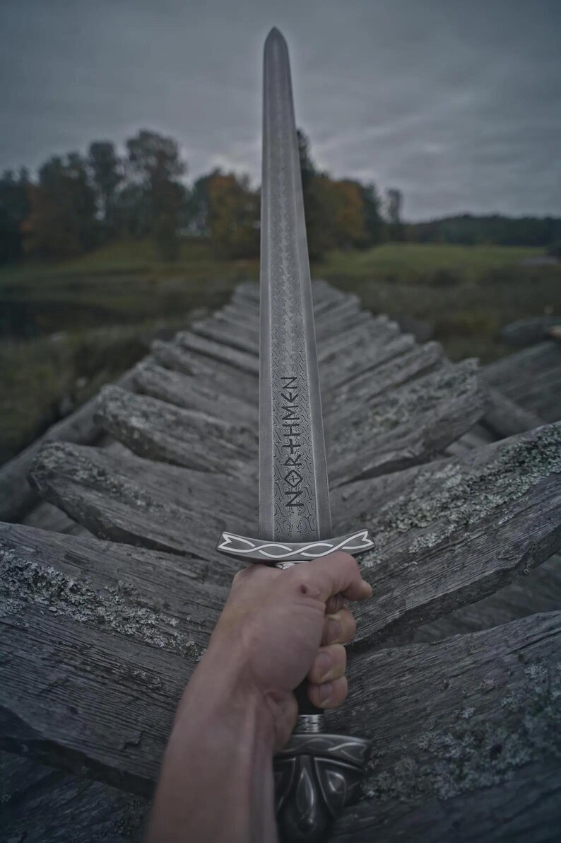 May include: A silver sword with the word "NORTHMEN" etched on the blade. The sword is held in a hand against a backdrop of weathered wooden planks and a blurred landscape under a cloudy sky. The sword's hilt has an intricate design.