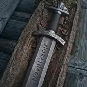 May include: A close-up of a Viking sword, with the word "NORTHMEN" inscribed on the blade. The sword has a silver-toned blade and hilt, and a black handle. It is displayed on a wooden surface, suggesting a historical or artisan context.