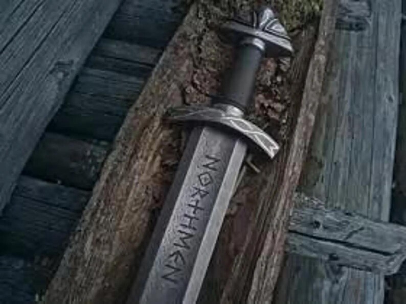 May include: A close-up of a Viking sword, with the word "NORTHMEN" inscribed on the blade. The sword has a silver-toned blade and hilt, and a black handle. It is displayed on a wooden surface, suggesting a historical or artisan context.