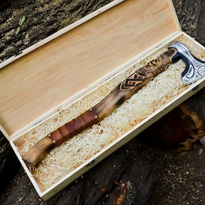 May include: A handcrafted axe with a wooden handle and a decorative metal head, presented in a wooden box. The handle features intricate carvings and a leather grip. The axe head has a silver and black design.