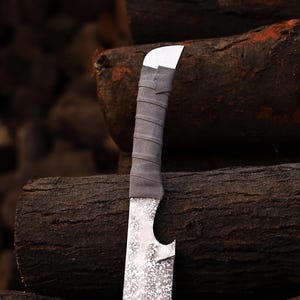 May include: A close-up shot of a handmade knife. It features a textured silver blade and a grey, wrapped handle. The knife has a curved blade and a white metal top. The background shows stacked logs.