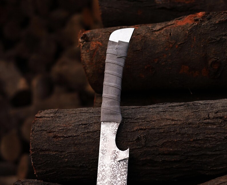 May include: A close-up shot of a handmade knife. It features a textured silver blade and a grey, wrapped handle. The knife has a curved blade and a white metal top. The background shows stacked logs.
