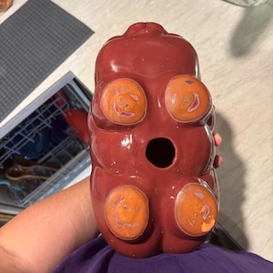 May include: A glossy, red ceramic bear-shaped container with four orange, circular accents. The bear has a large opening and is held by a person wearing a purple shirt. The container is likely a decorative item or a unique storage piece.