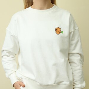Puede incluir: Sudadera blanca con un pequeño bordado de capibara de dibujos animados y una hoja verde. El capibara es marrón y naranja. La sudadera tiene cuello redondo y mangas largas. El estilo es casual y cómodo.