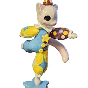 May include: A whimsical ceramic figurine of a white cat dressed as a circus clown. The cat balances on a star-shaped base, wearing a blue and yellow outfit with polka dots and a matching hat. The figurine has a playful, artistic design.