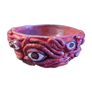 May include: A red, sculpted bowl with a textured exterior. The bowl features multiple eye designs around the rim, with white and brown details. The interior of the bowl is a lighter shade of red.