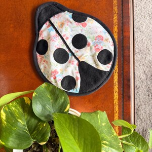 May include: A ladybug-shaped trivet with a black border and large black spots. The center features a floral pattern with pink, blue, and yellow flowers on a white background. The trivet is placed on a brown surface.