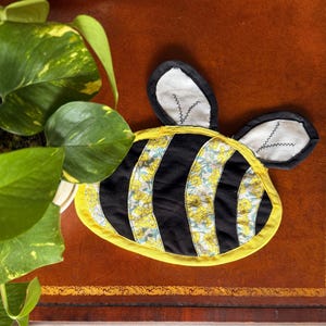 Könnte beinhalten: Handgefertigter Topfuntersetzer in Bienenform mit schwarzen und gelben Streifen und floralen Akzenten. Der Untersetzer hat einen gelben Rand und schwarz-weiße Ohren. Schützt Oberflächen vor Hitze und ist ein dekoratives Küchenaccessoire.