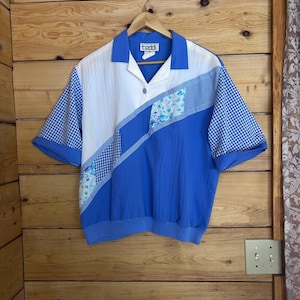 Puede incluir: Una camisa de manga corta con cuello y cierre de un solo botón. El diseño presenta un patrón de bloques de color blanco, azul sólido, cuadros vichy azules y blancos, y un panel con estampado floral. La camisa tiene una etiqueta que dice teddi.