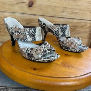 May include: A pair of open-toe high-heeled sandals with a snakeskin pattern in shades of brown, black, and beige. The sandals feature two straps with buckle closures and a platform sole. The heels are approximately 3 inches high.