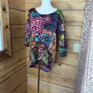 May include: A long-sleeved top featuring a textured, crinkled fabric with a multi-coloured patchwork pattern. The design includes vibrant shades of magenta, teal, lime green, and orange in various abstract shapes and floral-inspired motifs.