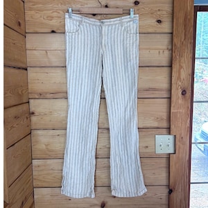 May include: Cream-coloured, vertically striped trousers with a flared leg. The trousers have a button and zip closure, belt loops, and front and back pockets. The trousers are hanging on a wooden hanger against a wooden wall.