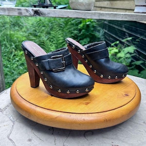 Può includere: Un paio di zoccoli in pelle nera con tacchi e plateau alti in legno. Ogni scarpa presenta un cinturino decorativo con fibbia in metallo sul collo del piede, borchie in metallo argentato lungo la base e il marchio ALDO sulla soletta.