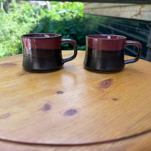 May include: Two ceramic mugs featuring a two-tone design with a glossy dark red upper section and a solid black lower section. Each mug has a wide, rounded body, a short handle on the side, and a smooth, reflective finish on the exterior surface.